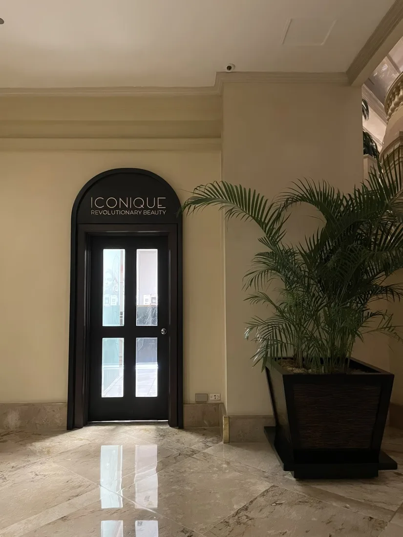 The ICONIQUE entrance: an arched dark wood door with the ICONIQUE sign, a palm and marble floor in the lobby of The Peninsula Manila